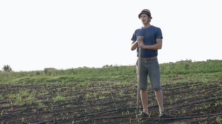 Potrait of caucasian farmer in hat with hoe is resting and while removes weeds in corn field at organic farm. Worker lean on the hoe and resting in weeding process.の写真素材