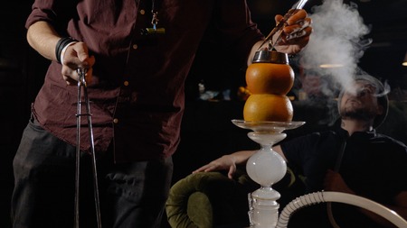 Young hookahman changing the coals in the hookah while men is smoking at dark shisha loungeの写真素材