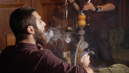 Young hookahman changing the coals in the hookah while men is smoking at dark shisha lounge and...