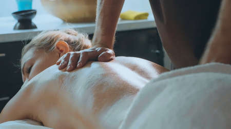 Beautiful blond girl lying on couch receiving back massage at luxury spa salon. Young woman relaxing and enjoying of professional massaje of back at luxury spa salonの写真素材