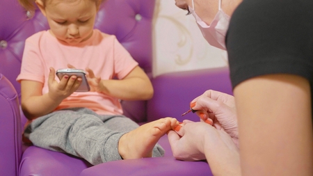 Cute baby girl using smart phone at pedicure procedure at beauty spa salon, close-up.の写真素材