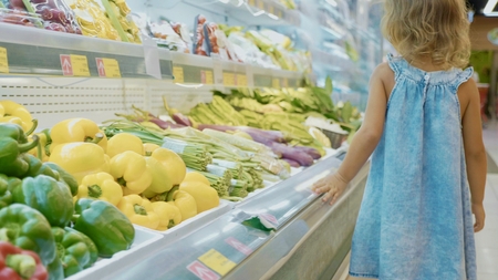 Little girl walking past the shelves with vegetables at grocery store, rear viewの写真素材