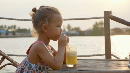 Funny little girl is sitting in a riverside cafe at a table and drinking juiceの写真素材