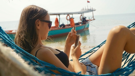 Young woman lying on hammock with smart phone and bank card at the sandy beach