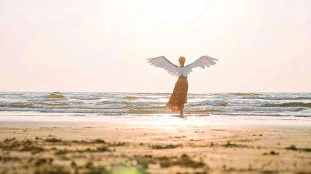 Beautiful female angel walking barefoot toward the sea at sunset.