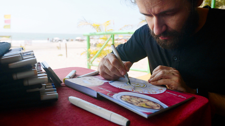 Man sits in cafe and drawing image of Indian food in sketchbook by art markers