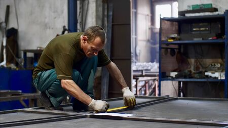 Man worker locksmith measures tape measure length of metal bars. He makes marks with pencil in...