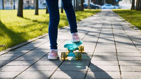Close-up back view of legs of a child girl in jeans and pink sneakers riding a skateboard in the park along the green grass in sunny weather.の写真素材