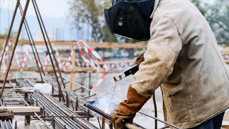 A worker at construction site is welding iron pipe with electrode in a protective uniform, orange gloves and black helmet. Sparks and smoke are allocated. Side view of an unrecognizable welder.の写真素材