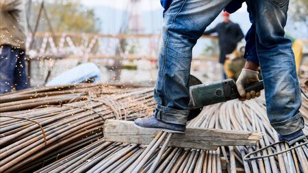 Close-up view of legs in foreground of a worker man in blue jeans, cutting rebar with a saw blade at construction site. Right leg is on the metal tubes which are on wooden stud, sparks from process.の写真素材