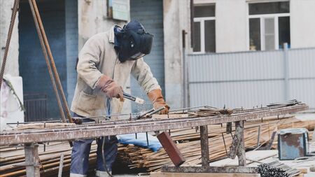 Welder at construction site is welding pipe with electrode in helmet.