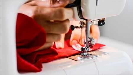 Hands of woman tailor sewing red clothing on sewing machine with straight seam.