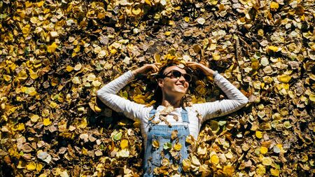 Cheerful young woman in denim overall is lying on autumnal yellow leaves
