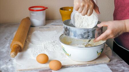 Mature woman is adding a flour through sieve in steel bowl for preparing a dough, close-up side view