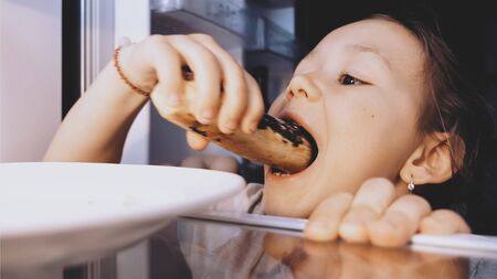 Child girl is opening a fridge, taking an eclair and eating it furtively.