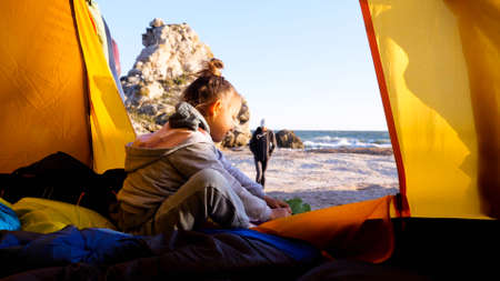 Child girl is puts her shoes while sits in camping tent at beautiful beachの写真素材