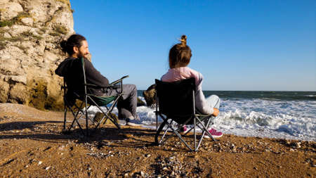 Father with little daughter sits on chairs on the beach, rear view.の写真素材
