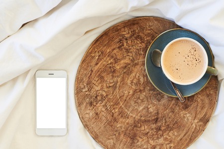White smartphone with blank copy space screen and a cup of coffee on a rustic wooden tray on the bedの写真素材