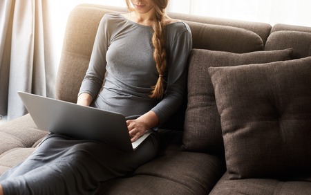 Beautiful young blond woman with braid working on a laptop sitting on the comfortable dark sofa at home, backlit warm light. Freelance or lifestyle concept. Empty space for your promotional contentの写真素材