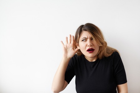 Portrait of funny plus size young woman looking aside holding hand to her ear and trying to hear something standing on white background. Cute Caucasian female with a hearing disorder or hearing lossの写真素材