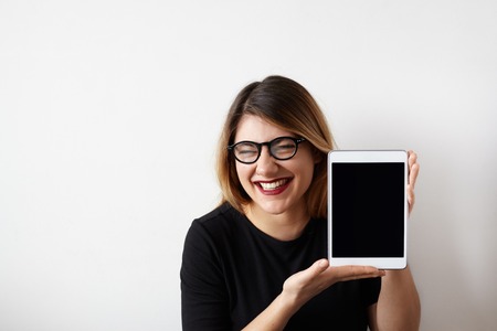 Humorous portrait of attractive young woman in glasses showing blank copy screen tablet, having funny sarcastic face expression, loud laughing meaning gloating, talking "muahaha". Copy space for textの写真素材