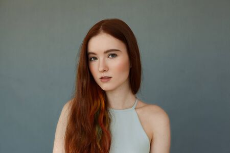 Portrait of young tender redhead teenage girl with green eyes wearing blue top looking at camera with serious or pensive expression standing on blue wall background, her lips slightly partedの写真素材