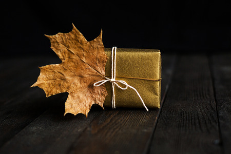 A golden gift box on black wooden background, decorated with dry maple leaves.Holiday concept, greeting card. Copy spaceの写真素材