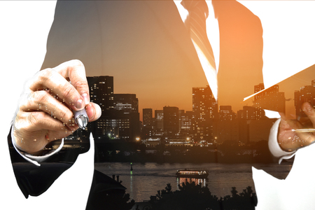 Businessman holding memo and ready to write down with big town or city in the background. Double exposure, panoramic view contemporary megalopolis background, orange sun light, business conceptの写真素材
