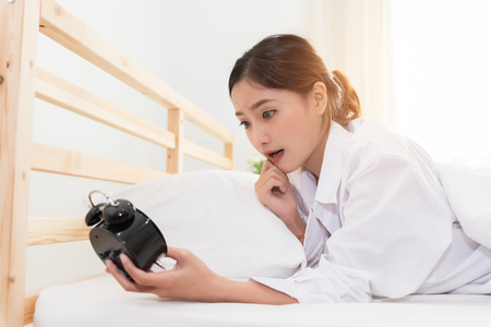 Asian woman shocked when wake up by alarm clock because too late for meeting appointment or working. Lazy day and Hurry up concept.の写真素材
