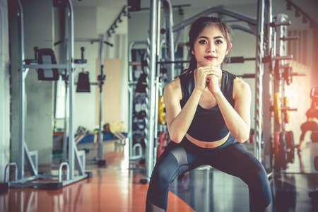 Young Asian woman doing squat workout for fat burning and diet in fitness sports gym with sports equipment in background. Beauty and body build up concept. Sports club and Aerobic theme.の写真素材