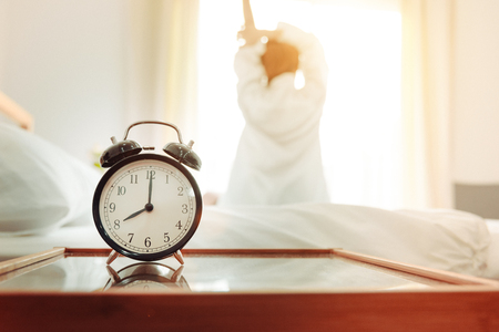 Back view of woman stretching in morning after waking up on bed near window with alarm clock. Holiday and Relax concept. Lazy day and Working day concept. Office woman and worker in daily life themeの写真素材