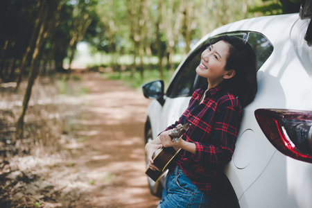 Beauty Asian woman smiling and having fun at outdoors summer with Ukulele near white car. Traveling of photographer concept. Hipster style and Solo woman theme. Lifestyle and Happiness life theme.の写真素材