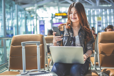 Young Asian female passenger using laptop and smart phone while sitting on seat in terminal hall and waiting for flight in airport . People lifestyles and Happy woman. Technology and Travel concept.の写真素材