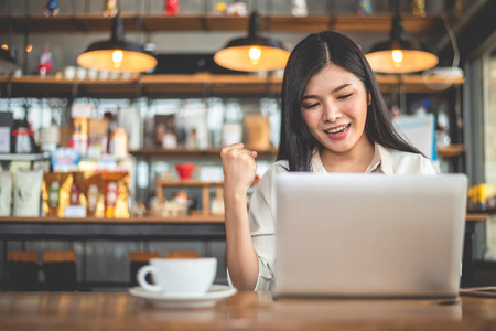 Asian female freelancer doing happy gesture by raising hand when using laptop in cafe. Business and success concept. Coffee shop and outdoors theme. Cafeteria background.の写真素材