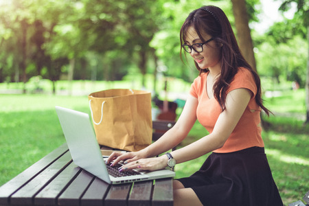 Asian woman using and typing on laptop keyboard in outdoors park. Woman chatting to her friends on social network. People and lifestyle concept. Nature and technology theme.の写真素材