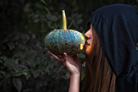 Female wizard kissing pumpkin lantern. Female costume in black clothes and hood on mystery forest...
