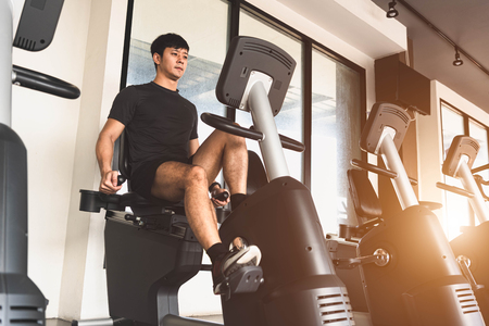 Asian young sport man riding stationary bicycle in fitness gym. Man working out on spinning bikes in gym. People lifestyles and health club concept. Bodybuilding themeの写真素材