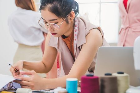 Attractive Asian female fashion designer working in clothing shop with customer background. Stylish fashionista woman creating new cloth design collection. Tailor and sewing. People lifestyle conceptの写真素材