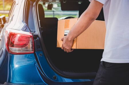 Delivery man is delivering cardboard box to customers via private car trunk door. People lifestyles and business occupation concept. Young male courier in casual clothes. Parcel move courier serviceの写真素材