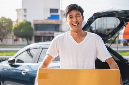Happy Asian man is delivering cardboard box to customers via private car. People lifestyles and business occupation concept. Young male courier in casual clothesの写真素材