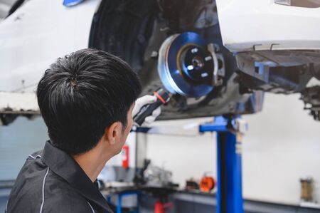Asian male mechanical hold and shining flashlight to examine car disk brake pad wear of automotive vehicle. Safety suspension inspection check service maintenance for customer before road trip conceptの写真素材