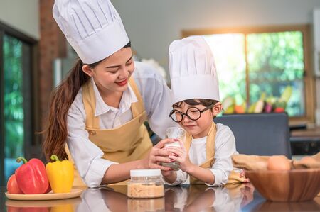 Beautiful Asian woman teaching cute little boy with eyeglasses to drink milk in kitchen at home together. Lifestyles and Family. Son dislike milk and doing funny face with milk glass. Food and drinkの写真素材