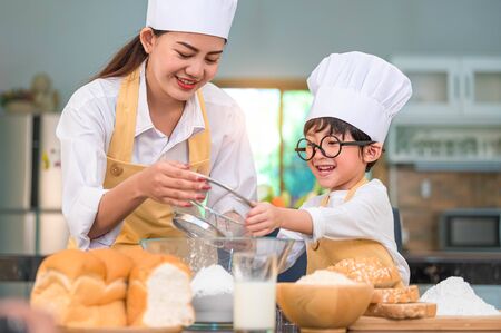 Cute little Asian boy and beautiful mother sifting dough flour with sifter sieve colander in home kitchen on table for prepare to baking bakery and cake. Thai kids playing with flour as chef funnyの写真素材
