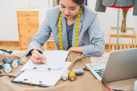 Attractive Asian female fashion designer working in home office workshop. Stylish fashionista woman creating new cloth design collection. Tailor and sewing. People lifestyle and occupation conceptの写真素材