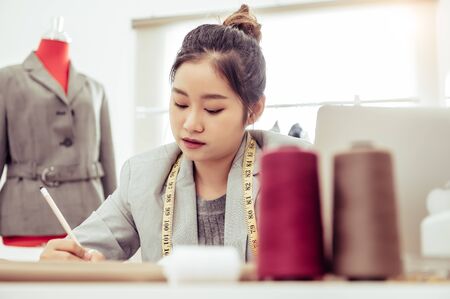 Attractive Asian female fashion designer working in home office workshop. Stylish fashionista woman creating new cloth design collection. Tailor and sewing. People lifestyle and occupation conceptの写真素材
