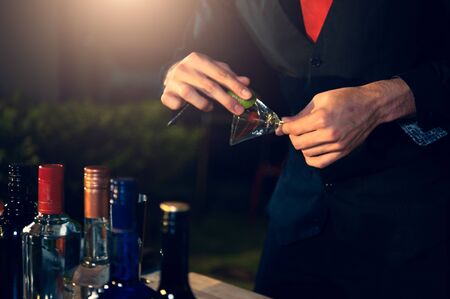 Professional bartender preparing fresh lime lemonade cocktail in drinking wine glass with ice at night bar clubbing counter. Occupation and people lifestyles concept. Outdoor backgroundの写真素材