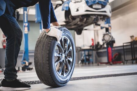 Male mechanic hold and rolling tire at repairing service garage background. Technician man replacing winter and summer tyre for safety road trip. Transportation and automotive maintenance conceptの写真素材