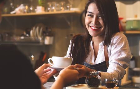 Beautiful Asian female barista serving hot coffee to customer at counter bar background. Smiling...