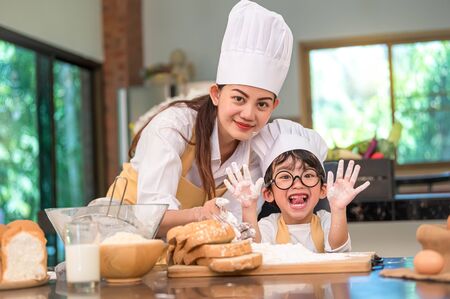 Beautiful woman and cute little Asian boy with eyeglasses, chef hat and apron playing and baking bakery in home kitchen funny concept. Homemade food and bread. Funny face emotion looking cameraの写真素材