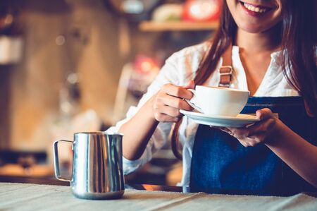 Closeup of professional female barista hand making and holding white cup of coffee. Happy young woman at counter bar in restaurant background. People lifestyles and Business occupation conceptの写真素材